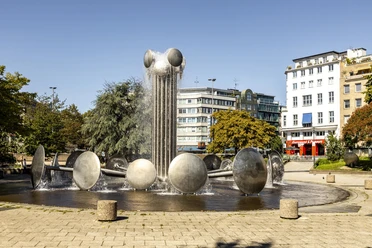 Fountain at Ebertplatz Ebertplatz in Cologne features a modern fountain with water drums and a central column. Surrounded by trees and buildings, it provides an urban backdrop on a sunny day.