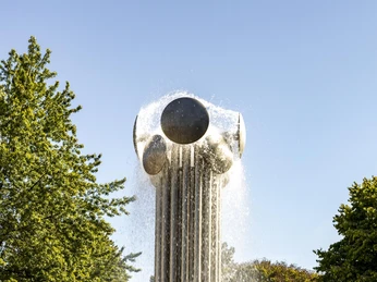 Ebertplatz Im Zentrum des Bildes erhebt sich eine große, futuristische Skulptur mit Wasserfontänen. Umgeben von kugelförmigen Elementen, schimmert der Brunnen im Sonnenlicht, eingerahmt von üppigem Grün der umliegenden Bäume.A large, futuristic sculpture with water fountains rises in the center of the picture. Surrounded by spherical elements, the fountain shimmers in the sunlight, framed by the lush green of the surrounding trees.