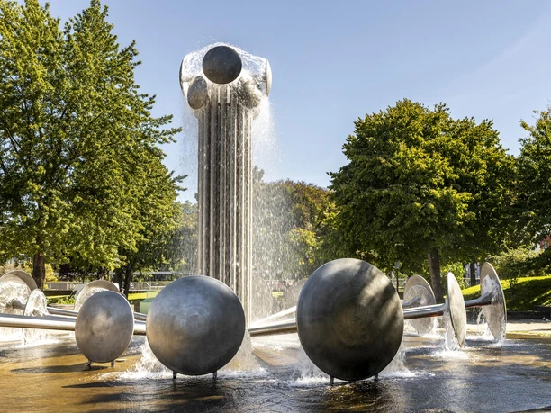 Ebertplatz A large, modern fountain on Ebertplatz in Cologne, surrounded by trees, in sunny weather.