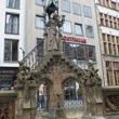 Steinbrunnen mit detailreicher Statue vor modernen und historischen Gebäuden in belebter Umgebung.