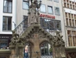 Steinbrunnen mit detailreicher Statue vor modernen und historischen Gebäuden in belebter Umgebung.