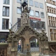 Steinbrunnen mit detailreicher Statue vor modernen und historischen Gebäuden in belebter Umgebung.
