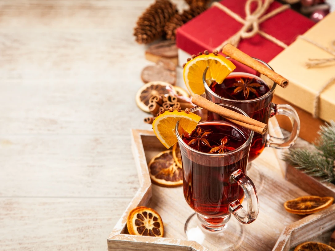 Zwei Gläser Glühwein mit Orangenscheiben und Zimtstangen, umgeben von Gewürzen und Weihnachtsdeko.