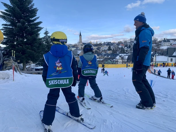 Eibenstock_Skiarena_Wurzelrudi_Winter©Wurzelrudi (9).jpg Zwei Kinder steigen im Skikurs mit Helm und Skilehrer den verschneiten Hang hinauf.