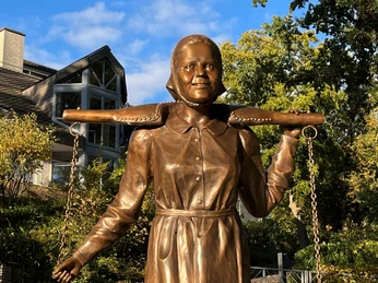 Die Melkerin Bronzestatue einer Frau in traditioneller Kleidung, die einen Melkstuhl auf den Schultern balanciert.