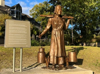 Die Melkerin Die Skulptur "Die Melkerin" aus Bronze zeigt eine Frau mit zwei Milchkannen über der Schulter.