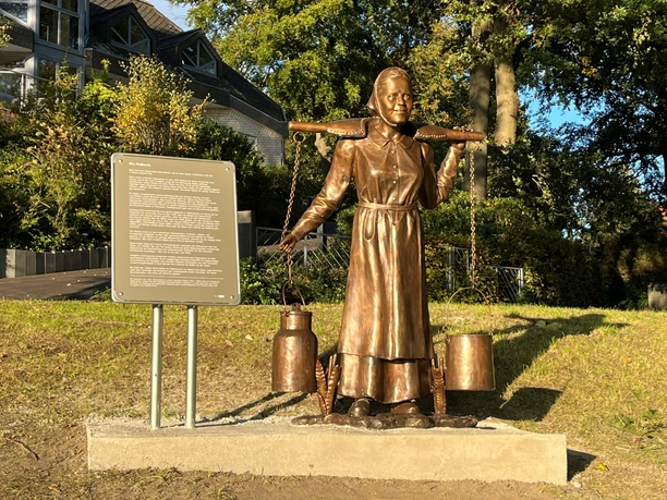 Bronzestatue einer Frau in traditioneller Kleidung, die zwei Milchkannen mit einer Tragevorrichtung trägt.
