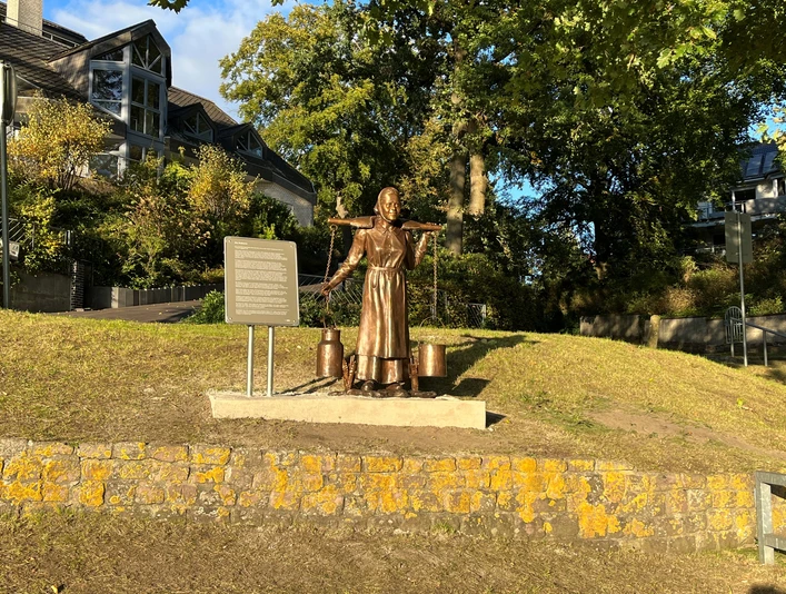 Eine Bronzeskulptur einer Frau mit einem Joch und Milchkannen steht vor einer Infotafel in einem Park.