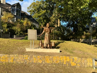 Die Melkerin Eine Bronzeskulptur einer Frau mit einem Joch und Milchkannen steht vor einer Infotafel in einem Park.