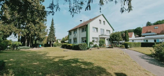 Frontansicht Zweistöckiges Wohnhaus im Grünen, von Bäumen umgeben, mit gepflegtem Garten und angrenzendem Weg.