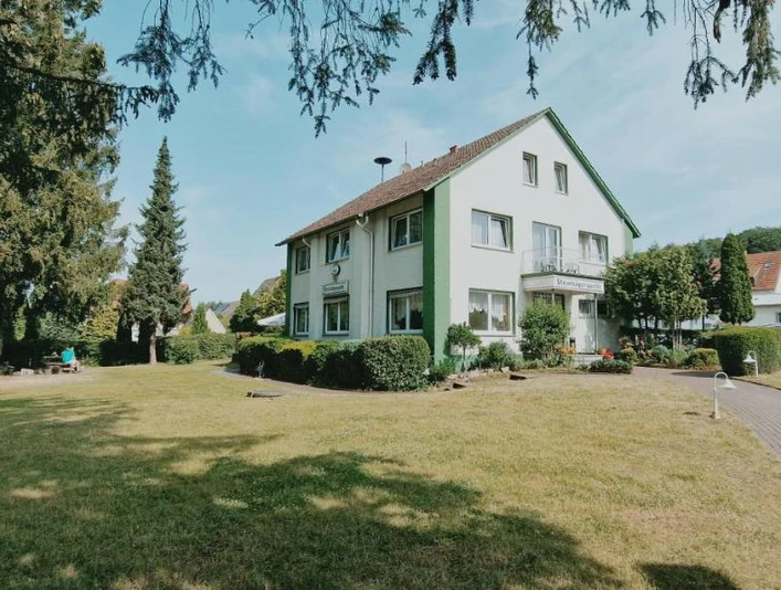 Frontansicht Zweistöckiges Wohnhaus im Grünen, von Bäumen umgeben, mit gepflegtem Garten und angrenzendem Weg.