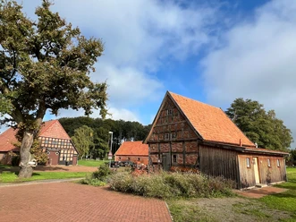 Noltesche Scheune Historische Fachwerkscheune mit rotem Dach in ländlicher Umgebung, von Bäumen umgeben, bei Sonnenschein.