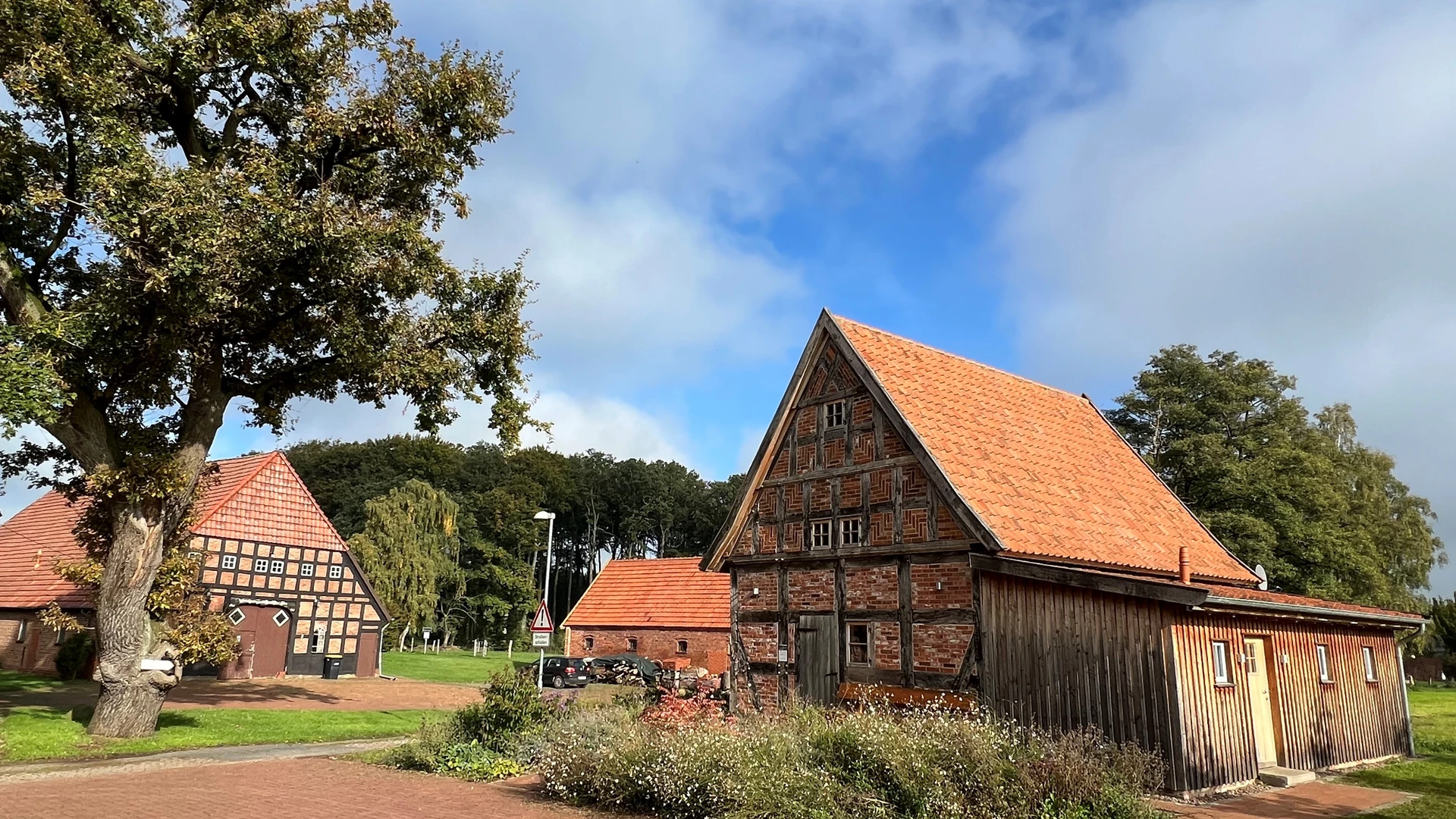 Historische Fachwerkscheune mit rotem Dach in ländlicher Umgebung, von Bäumen umgeben, bei Sonnenschein.