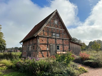 Eine historische Fachwerkscheune, umgeben von grünen Büschen und Blumen, unter einem teils bewölkten Himmel.