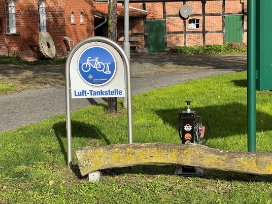 Grüne Wiese mit Luft-Tankstelle vor rotbraunen Backsteingebäuden und zwei runden Mühlsteinen.