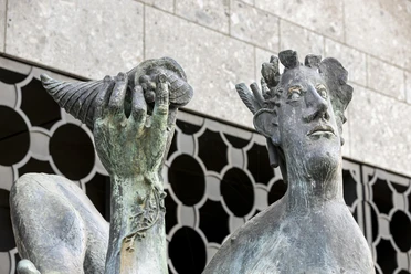 Dionysos Brunnen Bronzestatue des Dionysos an einem Brunnen in Köln, hält fröhlich eine Muschel in der Hand, kunstvoll gestaltet gegen modernen Steinbau. Die detaillierten Gesichtszüge und die mächtige Muschel verleihen dem Ensemble einen besonderen Charme.