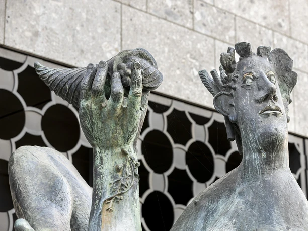 Dionysos Brunnen Bronzestatue des Dionysos an einem Brunnen in Köln, hält fröhlich eine Muschel in der Hand, kunstvoll gestaltet gegen modernen Steinbau. Die detaillierten Gesichtszüge und die mächtige Muschel verleihen dem Ensemble einen besonderen Charme.