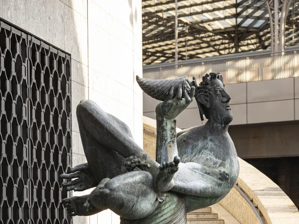 Dionysos Brunnen Die Skulptur des Dionysos-Brunnens in Köln zeigt eine dynamische Figur aufrecht, mit erhobenem Trinkkelch, auf einem runden Sockel stehend.