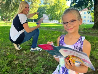 City Game Sebnitz Girl with glasses reads a colorful book in the park while a woman in the background smiles and points at something.