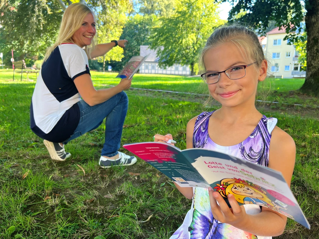 City Game Sebnitz Mädchen mit Brille liest ein buntes Buch im Park, während eine Frau im Hintergrund lächelnd auf etwas zeigt.Girl with glasses reads a colorful book in the park while a woman in the background smiles and points at something.Dívka s brýlemi si v parku čte barevnou knihu, zatímco žena v pozadí se usmívá a na něco ukazuje.Dziewczyna w okularach czyta kolorową książkę w parku, podczas gdy kobieta w tle uśmiecha się i wskazuje na coś.Meisje met bril leest een kleurrijk boek in het park, terwijl een vrouw op de achtergrond glimlacht en naar iets wijst.Una ragazza con gli occhiali legge un libro colorato nel parco, mentre una donna sullo sfondo sorride e indica qualcosa.