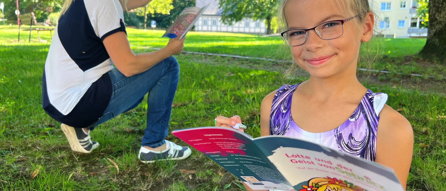City Game Sebnitz Girl with glasses reads a colorful book in the park while a woman in the background smiles and points at something.