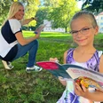 Stadtspiel Sebnitz Mädchen mit Brille liest ein buntes Buch im Park, während eine Frau im Hintergrund lächelnd auf etwas zeigt.Girl with glasses reads a colorful book in the park while a woman in the background smiles and points at something.Dívka s brýlemi si v parku čte barevnou knihu, zatímco žena v pozadí se usmívá a na něco ukazuje.Dziewczyna w okularach czyta kolorową książkę w parku, podczas gdy kobieta w tle uśmiecha się i wskazuje na coś.Meisje met bril leest een kleurrijk boek in het park, terwijl een vrouw op de achtergrond glimlacht en naar iets wijst.Una ragazza con gli occhiali legge un libro colorato nel parco, mentre una donna sullo sfondo sorride e indica qualcosa.