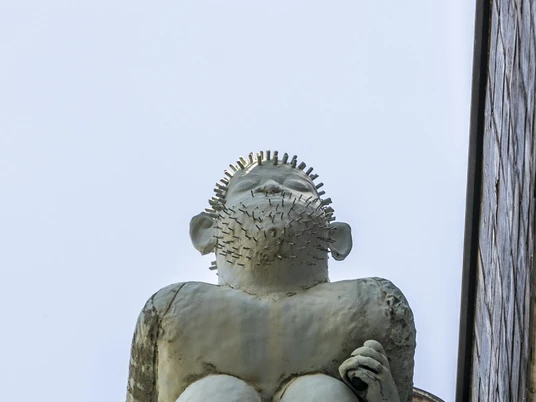 Kallendresser Das Bild zeigt die Skulptur "Kallendresser" auf einem Vorsprung an der Fassade, die eine Sitzfigur darstellt, die humorvoll die Hosen heruntergelassen hat. Die Figur hat stachelige Haare und richtet den Blick neugierig in die Ferne.The picture shows the sculpture "Kallendresser" on a projection on the façade, which depicts a seated figure that has humorously dropped its pants. The figure has spiky hair and looks curiously into the distance.