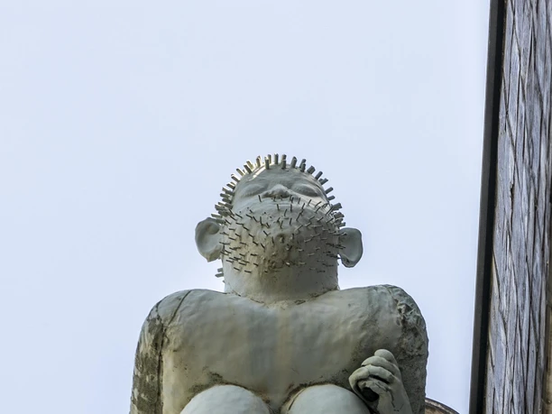 Kallendresser Das Bild zeigt die Skulptur "Kallendresser" auf einem Vorsprung an der Fassade, die eine Sitzfigur darstellt, die humorvoll die Hosen heruntergelassen hat. Die Figur hat stachelige Haare und richtet den Blick neugierig in die Ferne.