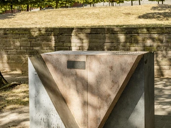 Rosa Winkel Memorial Denkmal aus geometrischen Betonformen im Schatten von Bäumen, mit modernem Gebäude im Hintergrund.Monument made of geometric concrete shapes in the shade of trees, with a modern building in the background.