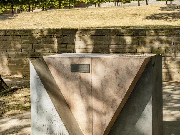 Rosa Winkel Memorial Monument made of geometric concrete shapes in the shade of trees, with a modern building in the background.