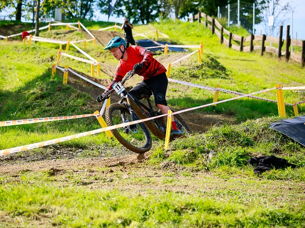 MTB-Rundkurs Ein Mountainbiker in rotem Trikot fährt dynamisch durch einen grasigen, abgesperrten Parcours.