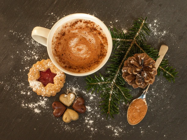 Eine Tasse heiße Schokolade, umgeben von Weihnachtsgebäck und Tannenzweigen auf dunklem Untergrund.