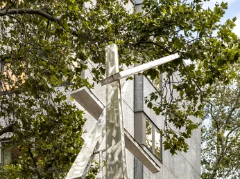 Four Trapezoids as two Rectangles IV Das Kunstwerk "Four Trapezoids as two Rectangles IV" zeigt zwei große, weiße Rechtecke, die an einem vertikalen Mast angebracht sind und sich scheinbar im Wind bewegen. Umrahmt von dichtem Laubwerk vor einem mehrstöckigen Gebäudefassaden-Hintergrund.The artwork "Four Trapezoids as two Rectangles IV" shows two large, white rectangles that are attached to a vertical pole and appear to move in the wind. They are framed by dense foliage in front of a multi-storey building façade background.