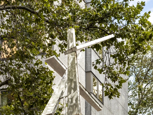 Four Trapezoids as two Rectangles IV The artwork "Four Trapezoids as two Rectangles IV" shows two large, white rectangles that are attached to a vertical pole and appear to move in the wind. They are framed by dense foliage in front of a multi-storey building façade background.