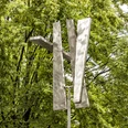 Four Trapezoids as two Rectangles IV Eine moderne Skulptur aus Aluminium-Trapezen erhebt sich vor dichtem grünen Laub.A modern sculpture made of aluminum trapezoids rises up in front of dense green foliage.