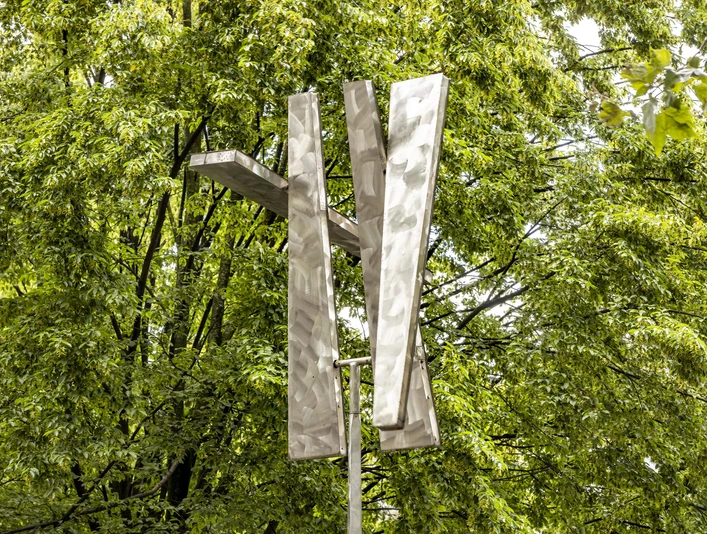 Four Trapezoids as two Rectangles IV Eine moderne Skulptur aus Aluminium-Trapezen erhebt sich vor dichtem grünen Laub.A modern sculpture made of aluminum trapezoids rises up in front of dense green foliage.