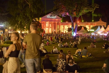 Parklichter Samstag Im Hintergrund ist die beleuchtete Wandelhalle zu sehen. Im Vordergrund versammeln sich einige Menschengruppen, während andere entspannt auf der Kurparkwiese verweilen.