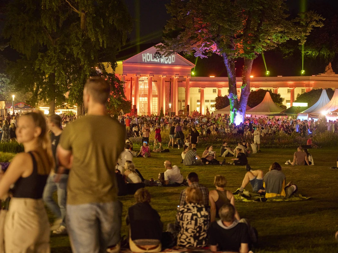 Parklichter Samstag Im Hintergrund ist die beleuchtete Wandelhalle zu sehen. Im Vordergrund versammeln sich einige Menschengruppen, während andere entspannt auf der Kurparkwiese verweilen.