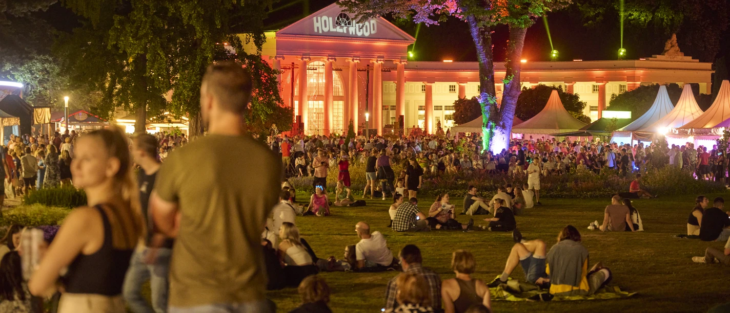 Parklichter Samstag Im Hintergrund ist die beleuchtete Wandelhalle zu sehen. Im Vordergrund versammeln sich einige Menschengruppen, während andere entspannt auf der Kurparkwiese verweilen.