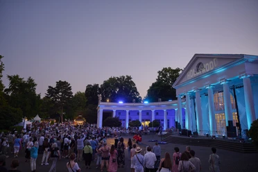 Parklichter Samstag Wandelhalle Rechts auf dem Bild erstrahlt die beleuchtete Wandelhalle im sanften Licht eines wunderschönen Sonnenuntergangs. Die belebten Menschengruppen verleihen der Szenerie eine lebendige Atmosphäre, während der warme Sommerabend eine unvergessliche Stimmung schafft.