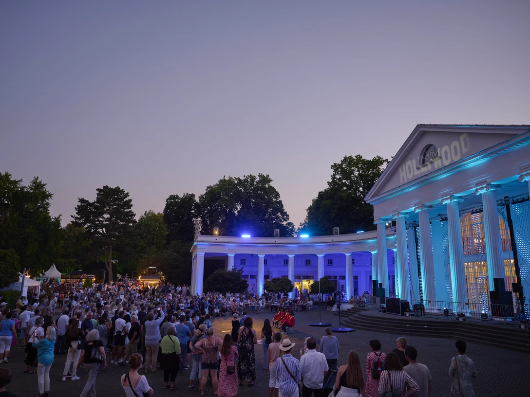 Parklichter Samstag Wandelhalle Rechts auf dem Bild erstrahlt die beleuchtete Wandelhalle im sanften Licht eines wunderschönen Sonnenuntergangs. Die belebten Menschengruppen verleihen der Szenerie eine lebendige Atmosphäre, während der warme Sommerabend eine unvergessliche Stimmung schafft.