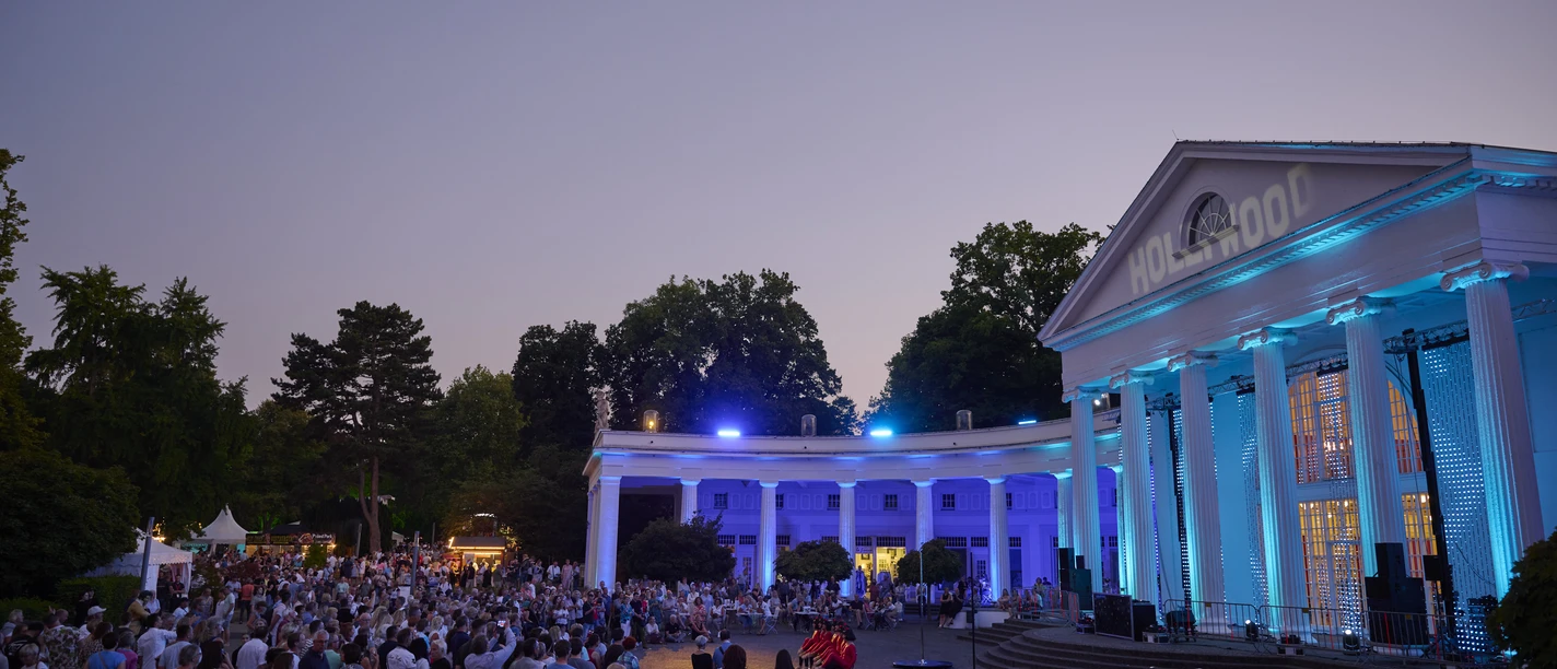 Parklichter Samstag Wandelhalle Rechts auf dem Bild erstrahlt die beleuchtete Wandelhalle im sanften Licht eines wunderschönen Sonnenuntergangs. Die belebten Menschengruppen verleihen der Szenerie eine lebendige Atmosphäre, während der warme Sommerabend eine unvergessliche Stimmung schafft.