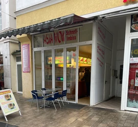 Das Bild zeigt einen Asia Wok Schnell-Imbiss mit Glasfronten und einem Tisch mit Stühlen davor.