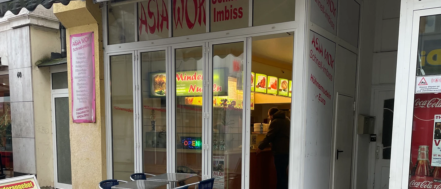 Das Bild zeigt einen Asia Wok Schnell-Imbiss mit Glasfronten und einem Tisch mit Stühlen davor.
