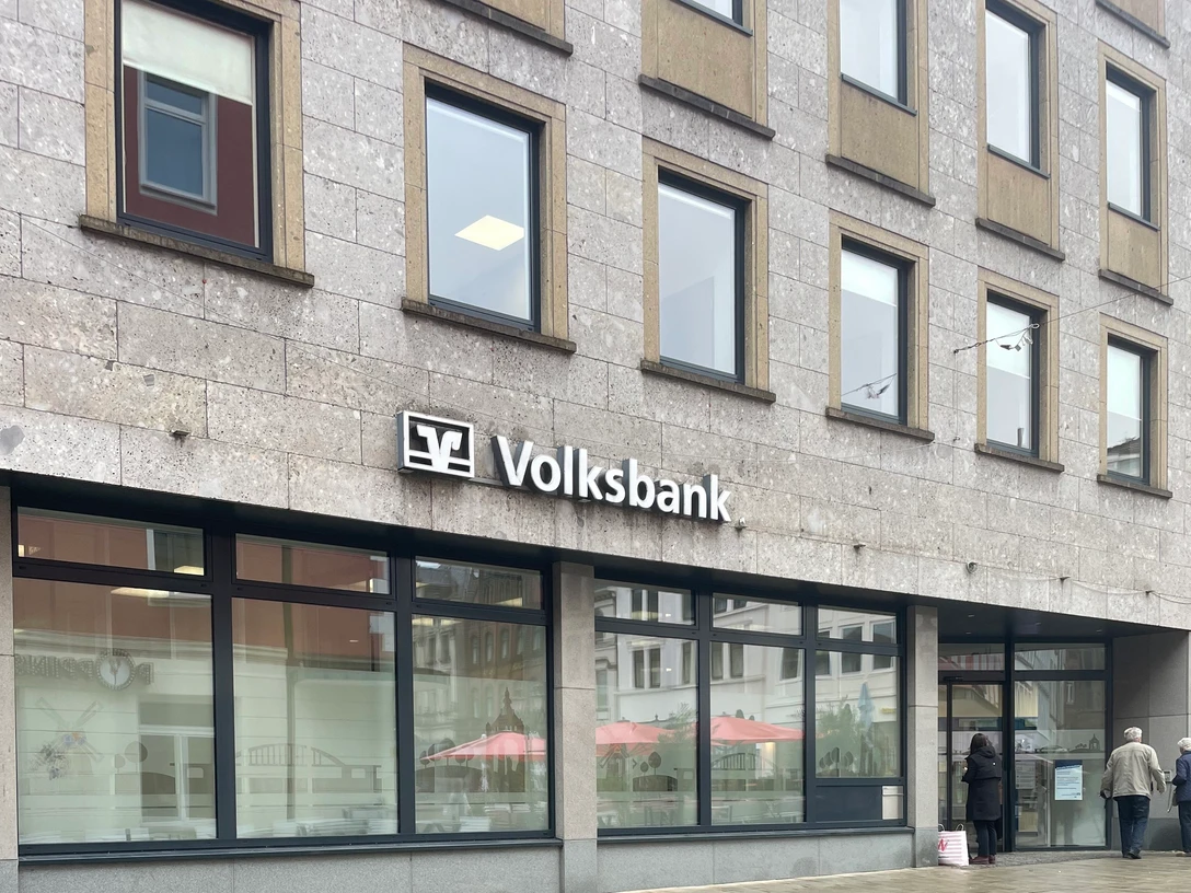 Eine Volksbank mit großem Logo auf grauer Fassade, großen Fenstern und Passanten davor.