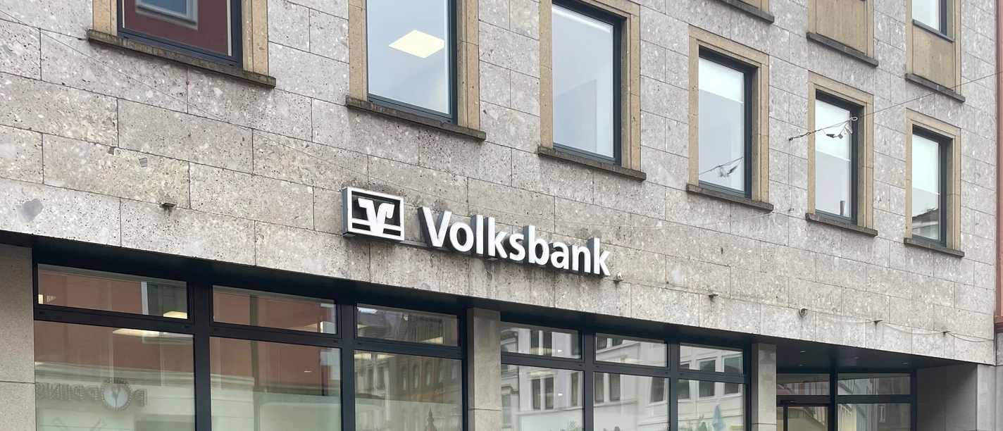 Voksbank.jpg Eine Volksbank mit großem Logo auf grauer Fassade, großen Fenstern und Passanten davor.
