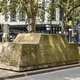 Ruhender Verkehr Ein moosbedeckter Betonblock mit Bäumen darauf steht inmitten einer urbanen Straßenszene.A moss-covered concrete block with trees on it stands in the middle of an urban street scene.