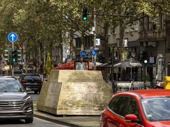 Ruhender Verkehr Am Bild sind parkende und fahrende Autos in einer belebten Kölner Straße zu sehen, flankiert von Geschäften und Bäumen. Ein massiver Betonblock in der Straßenmitte hebt sich inmitten des urbanen Verkehrs hervor, während die Ampeln den fließenden Verkehr steuern.The picture shows parked and moving cars in a busy street in Cologne, flanked by stores and trees. A massive concrete block in the middle of the street stands out amidst the urban traffic, while the traffic lights control the flow of traffic.