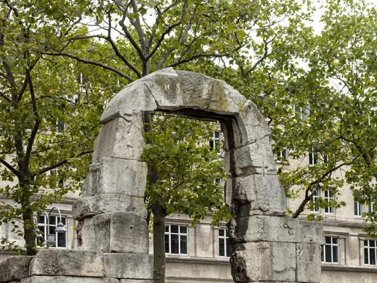 Römertorbogen Restaurierter Torbogen aus der römischen Antike in Köln, umrahmt von Bäumen, vor modernem Gebäude.Restored archway from Roman antiquity in Cologne, framed by trees, in front of a modern building.