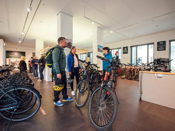 Tourist Service Bad Schandau Menschen bei Fahrradverleih, umgeben von Fahrrädern; ein Mann und eine Frau sprechen mit einem Mitarbeiter, während andere Gäste im Hintergrund stehen.